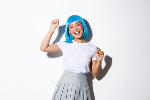 Image of joyful asian girl in blue wig dancing, celebrating halloween, standing happy over white background.