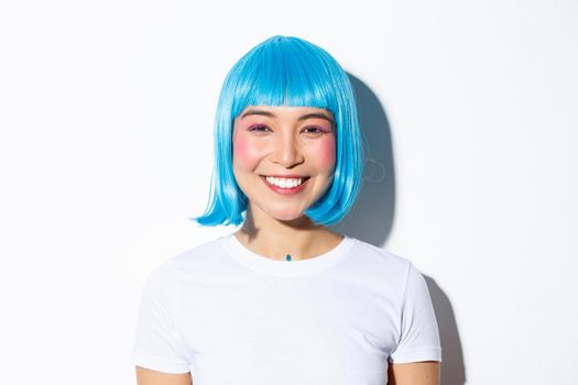 Close-up of cute asian female celebrating halloween in blue wig, smiling happy at camera, standing over white background.