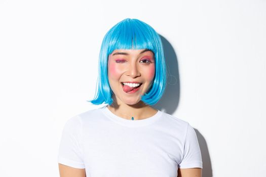 Close-up of attractive party girl in blue wig, showing tongue and winking happy, celebrating halloween, standing over white background.