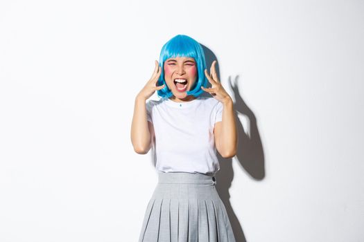 Portrait of distressed mad asian girl in blue wig screaming angry, standing over white background.