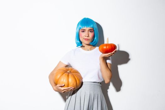 Image of indecisive cute asian girl in blue wig holding two different pumpkins and looking puzzled, standing over white background.