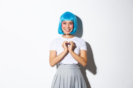 Image of lovely asian girl dressed-up in party costume, wearing wig and showing heart gesture, smiling at camera, standing over white background.