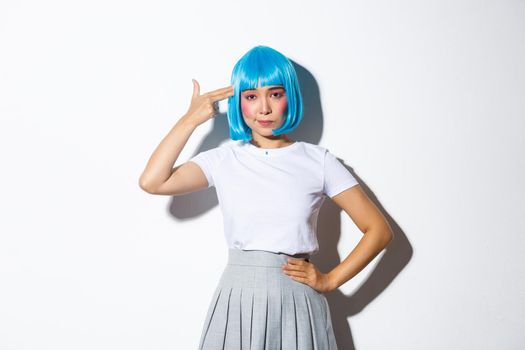 Image of bothered asian woman looking unamused while pointing hand gun at her head, wearing blue wig for halloween party, standing over white background.