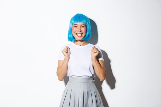 Image of cheerful asian girl feeling like winner, wearing blue wig, celebrating something, smiling and triumphing, standing over white background.