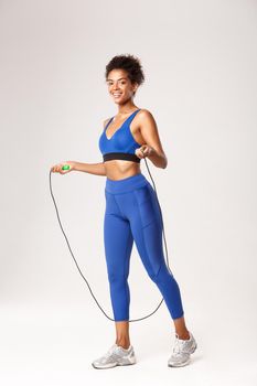 Full length of attractive fit african-american sportswoman, wearing blue uniform, jumping with skipping rope and smiling at camera, workoug against white background.