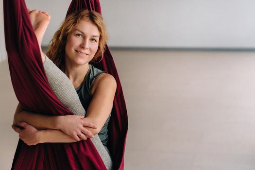 A woman does yoga sitting in a hanging hammock in the gym.