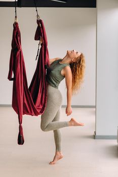 A happy woman does yoga on a suspended burgundy hammock in a bright gym.