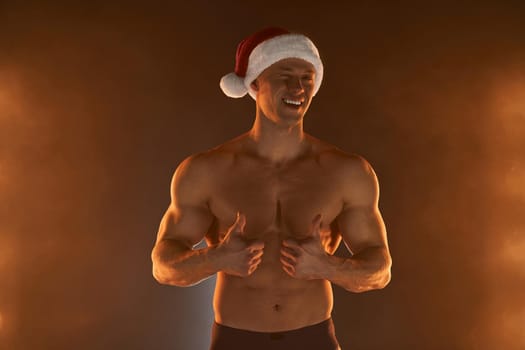 Portrait of muscular man wearing Christmas Santa hat, showing thumb up and smile on smoky background Macho Shirtless Naked torso stripper Hot sexy Santa guy Seductive male for holiday
