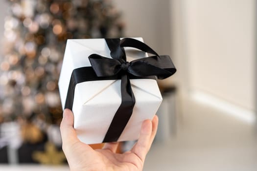 Man holding white Christmas gift box with black ribbon in hand with Christmas tree on background Surprise present box winter holiday