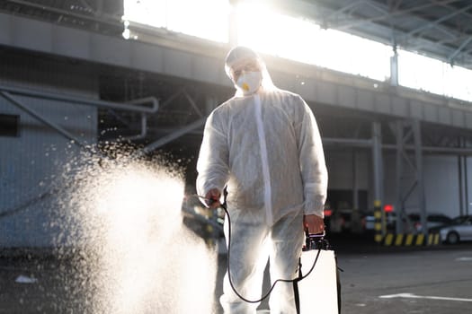 Man dressed white protective overalls spraying surface antibacterial sanitizer sprayer during quarantine Caucasian person protective suit disinfects parking during covid pandemic backlight