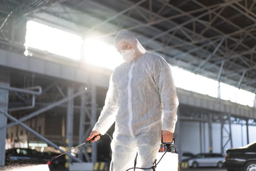 Man dressed white protective overalls spraying surface antibacterial sanitizer sprayer during quarantine Caucasian person protective suit disinfects parking during covid pandemic backlight