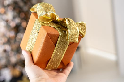 Man holding orange Christmas gift box with gold ribbon in hand with Christmas tree on background Surprise present box winter holiday