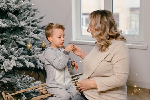 Holiday Christmas Caucasian mother playing with little cute son near decorated New year tree at home Family tradition