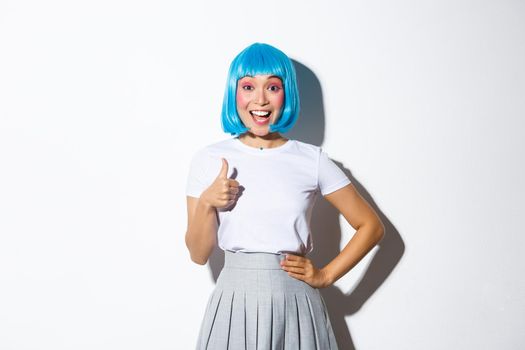 Portrait of excited smiling asian girl in blue party wig, showing thumb-up in approval, like something, standing in halloween costume over white background.