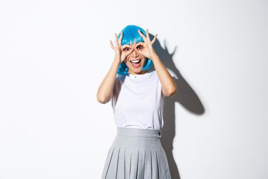 Portrait of cheerful asian girl looking amazed at camera, wearing blue wig and costume for halloween party, standing over white background.