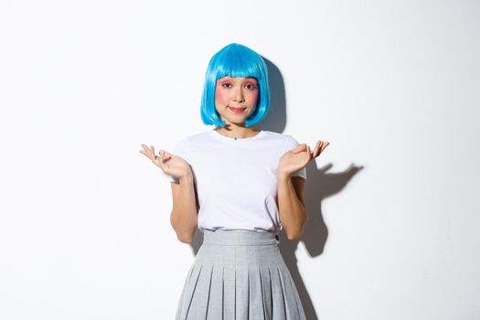 Portrait of clueless young asian party girl in blue wig shrugging, looking indecisive at camera, standing over white background.
