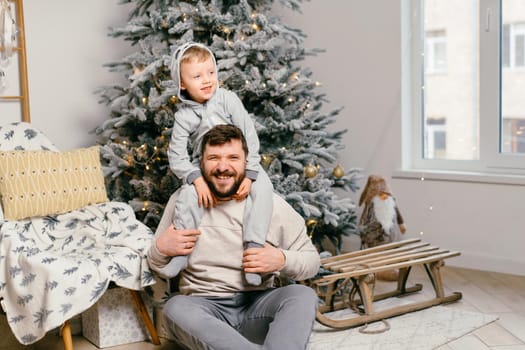 Holiday Christmas Handsome father playing with small cute little boy near decorated New year tree at home Family tradition positive son sitting on dad shoulders