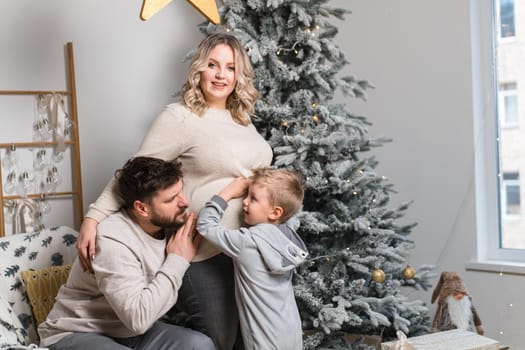 Christmas Family Happiness Portrait of dad, pregnant mom and little son sitting armchair at home near Christmas tree hug smile European young adult family holiday morning