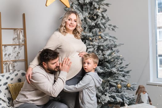 Christmas Family Happiness Portrait of dad, pregnant mom and little son sitting armchair at home near Christmas tree hug smile European young adult family holiday morning