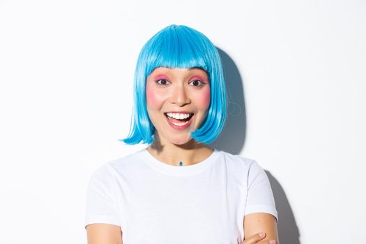 Image of excited cute asian girl in halloween costume and blue wig, smiling amused, standing over white background.
