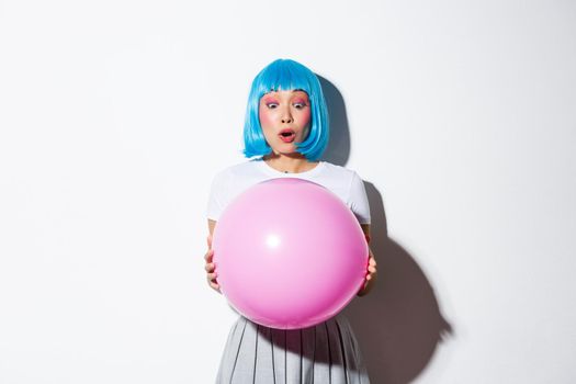Image of cheerful asian girl in blue wig, celebrating holiday, wearing outfit for halloween party, holding large pink balloon and looking amazed.