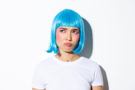 Image of cute asian girl in blue wig looking disappointed or jealous, sulking while stare at upper left corner, standing against white background.