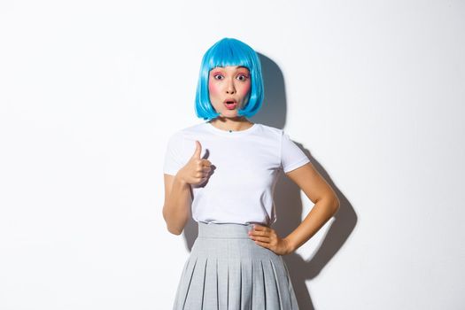 Amazed asian girl in blue party wig, showing thumbs-up in approval, looking impressed at camera, standing over white background.