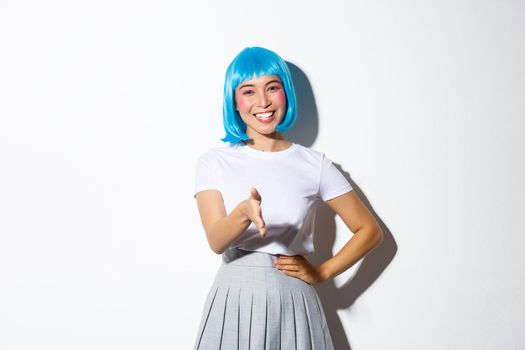Friendly cute asian girl in blue wig greeting someone with pleasant smile, giving handshake and saying hello, standing over white background in halloween costume.