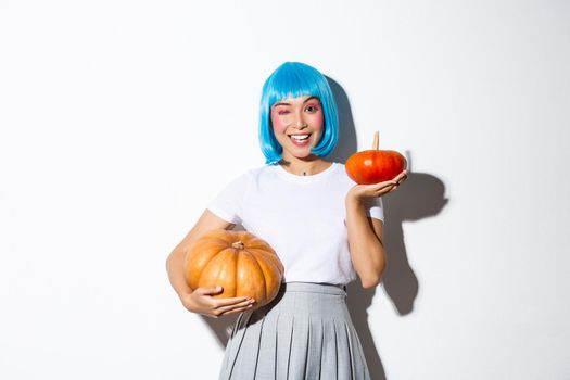 Happy beautiful asian girl celebrating halloween in blue wig and schoolgirl costume, winking at camera, showing big and small pumpkins.
