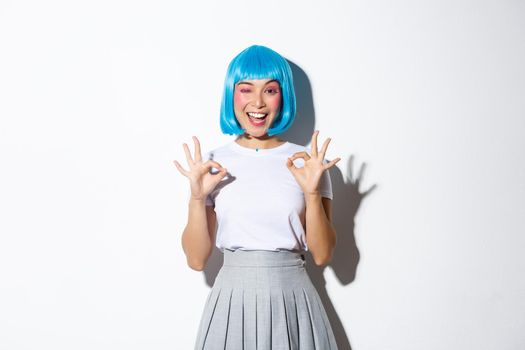 Image of happy smiling asian girl showing okay gesture and winking satisfied, standing in halloween costume over white background.