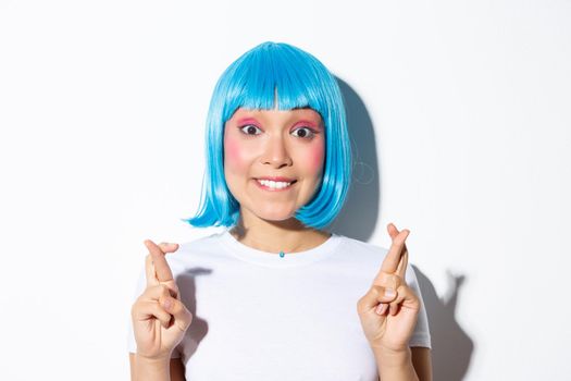 Excited beautiful asian girl in blue wig biting lip and looking hopeful at camera, making wish with fingers crossed, standing in halloween costume over white background.
