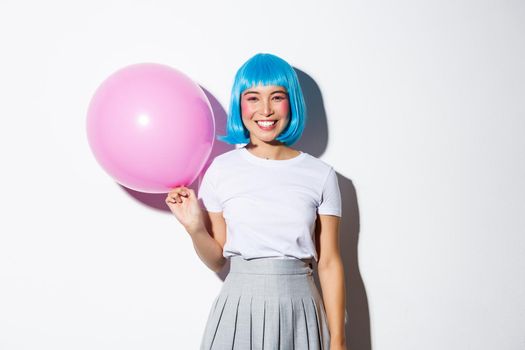 Cute asian female in blue wig and schoolgirl costume for halloween, holding pink balloon and smiling, standing over white background.