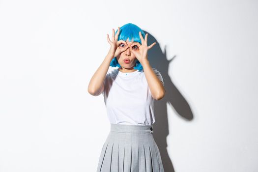 Portrait of amused beautiful asian girl in blue wig, looking through hand glasses with amazement, standing over white background.