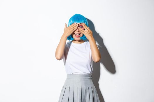 Excited beautiful asian girl peeking at camera amazed, looking at something interesting, dressed in blue wig and halloween costume, standing over white background.