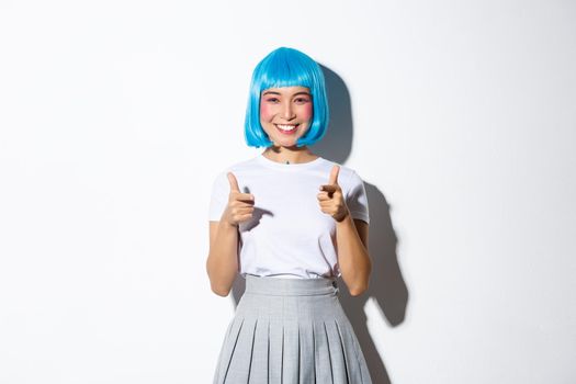 Cheeky japanese girl in blue party wig pointing fingers at camera and smiling, celebrating halloween, standing over white background.