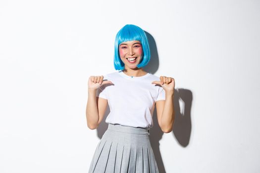 Happy asian girl in blue wig smiling and pointing at herself, celebrating halloween, standing over white background.