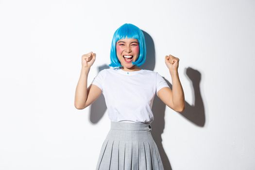 Portrait of successful happy girl in blue wig looking like winner, raising hands up and laughing, standing over white background.