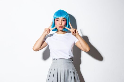 Cute asian pop girl in blue hair wig, pouting for kiss and showing kawaii peace gestures, wearing halloween party outfit, standing over white background.