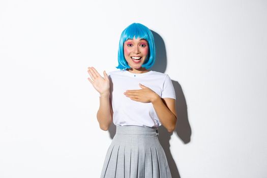 Portrait of cute asian girl in blue wig and halloween costume, smiling and looking sincere, holding hand on heart and making promise, standing over white background.