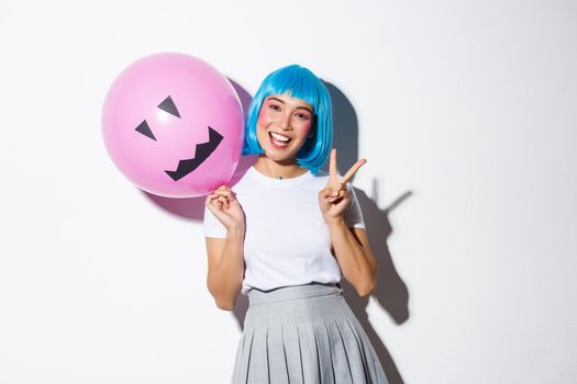 Image of happy asian girl celebrating halloween in blue anime wig, holding pink balloon with scary face and showing peace gesture.