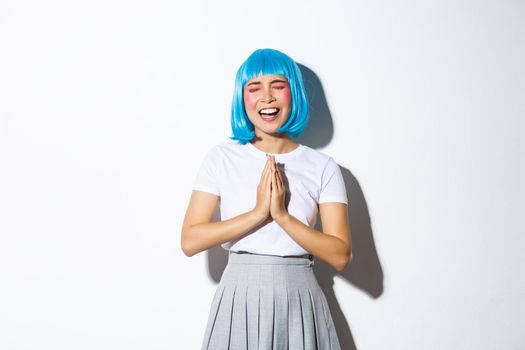 Portrait of silly asian girl in blue wig begging for help, pleading while standing over white background.