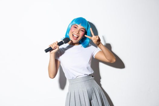 Cheerful cute asian girl dressed up as anime character for halloween party, wearing blue wig, holding microphone and singing karaoke, showing peace gesture.