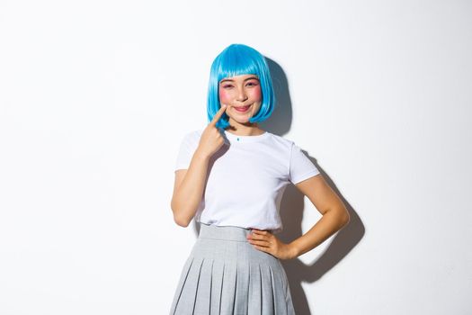 Image of kawaii asian girl in blue wig, smiling and pointing at her dimples, standing over white background, dressed for halloween party.
