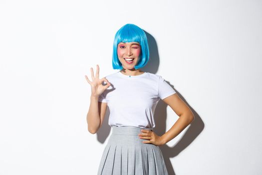 Portrait of confident attractive asian girl in blue short wig winking and showing okay gesture, dressed up for party or halloween celebration, standing over white background.