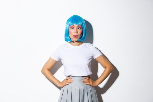 Portrait of silly asian girl in blue wig and party outfit showing tongue, squinting and making funny faces, standing over white background in halloween outfit.