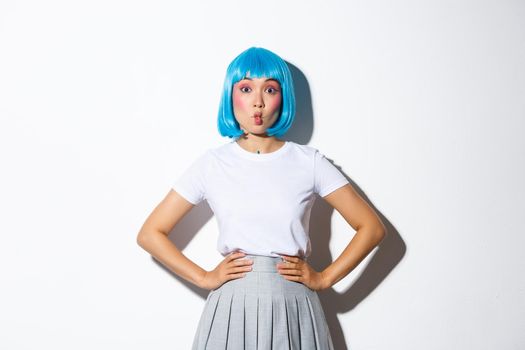 Silly asian girl in blue party wig, grimacing, making funny faces, wearing outfit for halloween or celebration events, standing over white background.