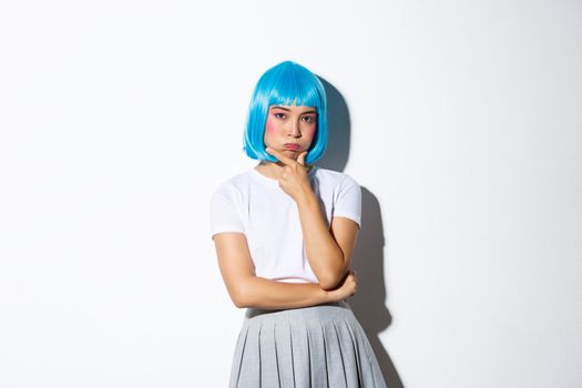 Portrait of troubled asian girl in blue wig pouting, looking complicated while pondering something, standing over white background.