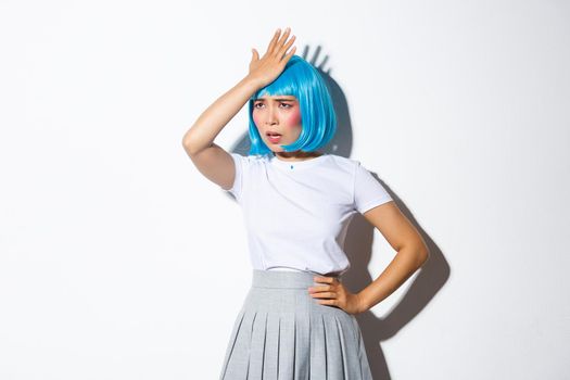 Portrait of bothered asian girl in blue party wig, slap forehead and looking pissed-off, forgot something, standing in halloween costume over white background.