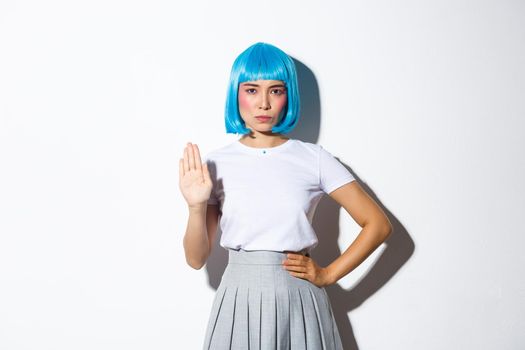 Portrait of serious and confident asian woman in blue wig showing stop gesture, disapprove or disagree with someone, standing over white background.