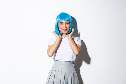 Portrait of beautiful asian girl in schoolgirl costume and blue wig, smiling kawaii at camera, standing over white background.